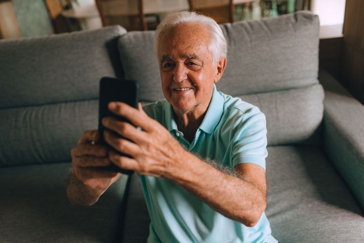 An older man sits on a sofa at home, smiling as he holds up his smartphone to check the readings he has just taken.