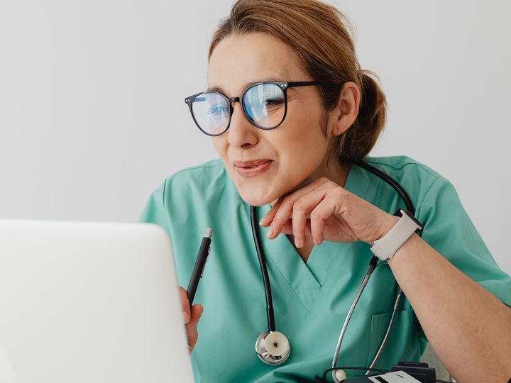 A clinician in scrubs, with a stethoscope around her neck, sits at a desk looking at an open laptop and holding a pen.