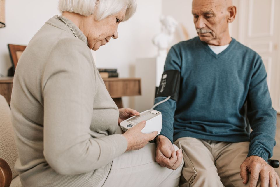 An older woman sits beside an older man in their living room, holding a digital blood pressure monitor and reading the display while he wears the inflated cuff on his upper arm.