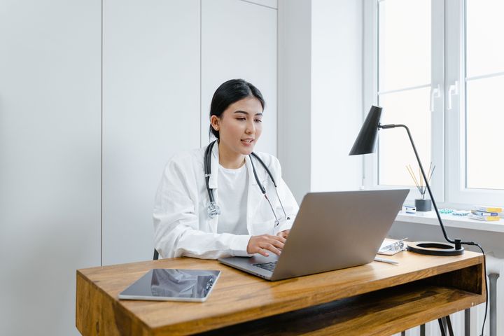 A clinician in a white coat with a stethoscope around her neck sits at a wooden desk by a window, typing on a laptop with a tablet beside her.