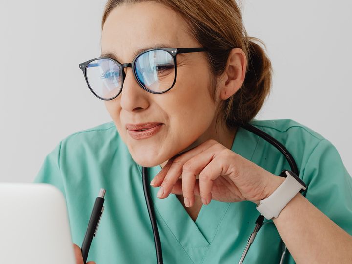A clinician in scrubs, with a stethoscope around her neck and a pen in hand, looking at a laptop screen.